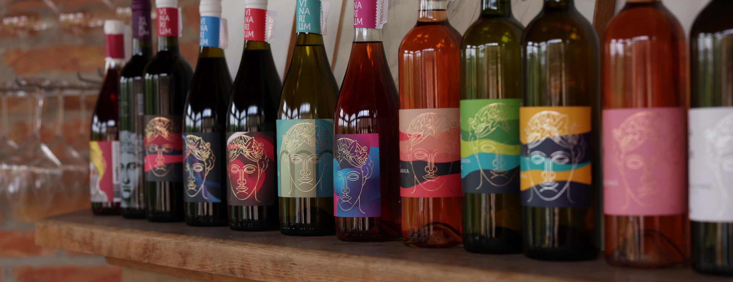 Slika 12 različnih steklenic rdečega, rožnatega in belega vina, ki stojijo na polici. Vse steklenice imajo barvito etiketo, na kateri je narisan obraz z lovorjevim vencem na glavi.