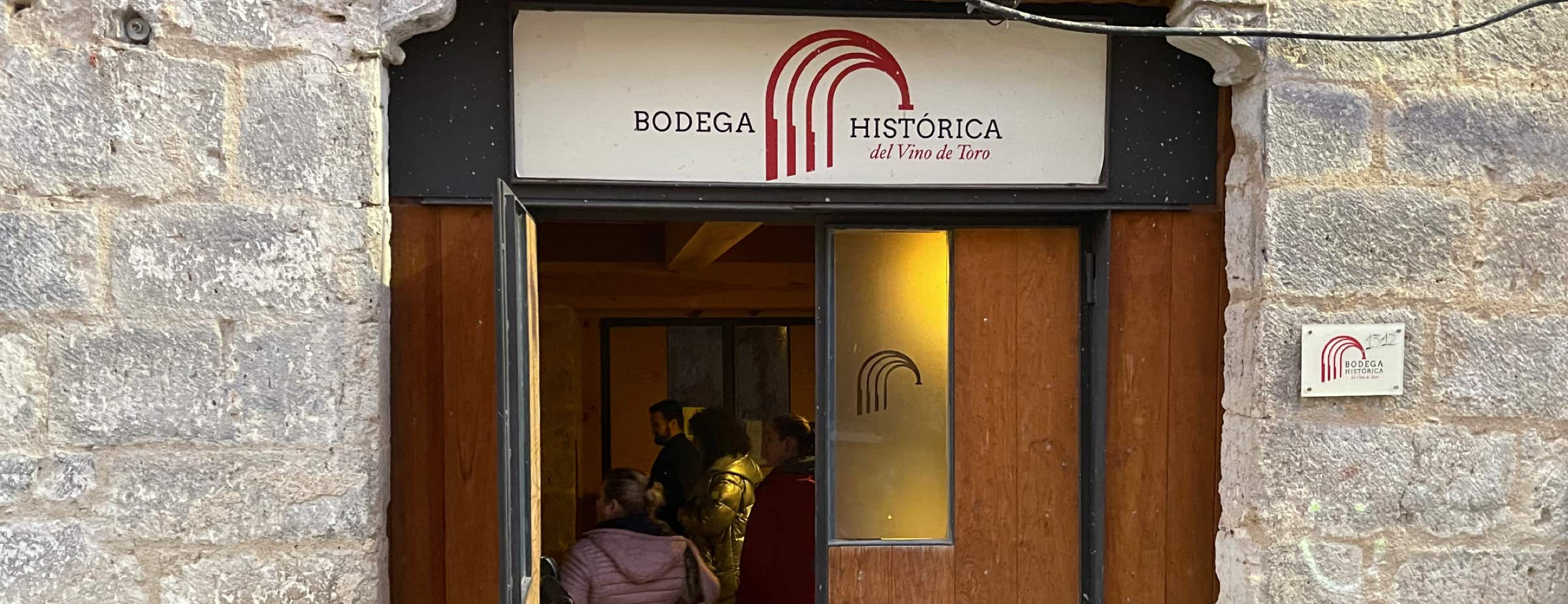 Picture of the entrance of the Bodega Histórica. The entrance is a wooden door. Over the entrance is a sign with “Bodega Histórica del Vino de Toro”. The door is open, and people can be seen inside. Next to the door, parts of the grey stone facade are visible.