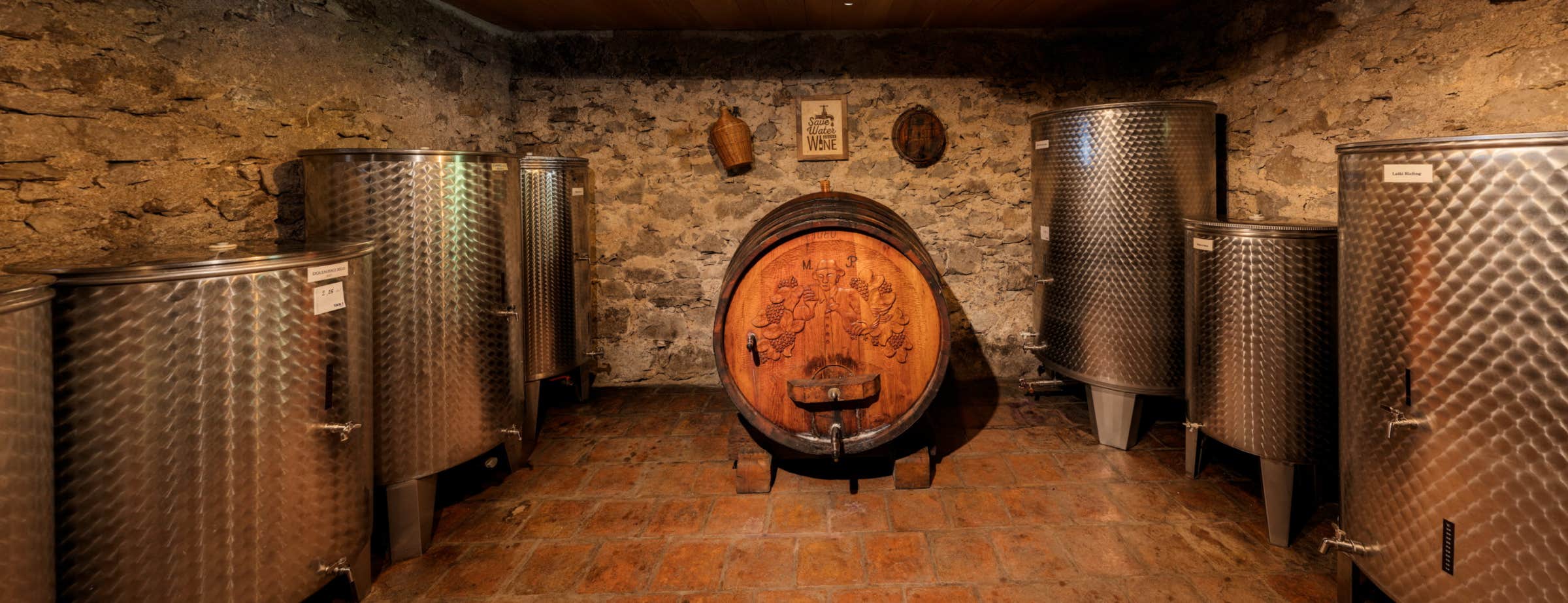 Image of a wine cellar showing a big wooden barrel with carvings of a man and grapes in front of the frontal wall in the middle of the picture. On the right and left side walls, multiple metal barrels are placed.