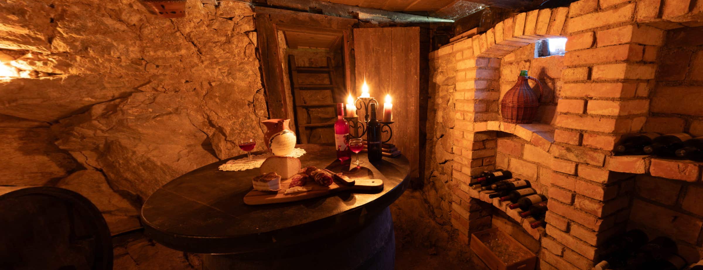 Image in wine cellar used for tastings. On a round table, wine, glassed, cheese, sausages and candles are decorated. On the right wall, stone shelfs are embedded into the wall and bottles of wine are lying on the shelfs.  