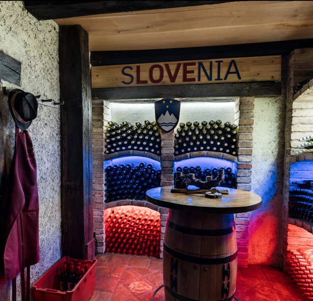 Dieses runde Bild zeigt einen Keller mit vielen Weinflaschen in einem Regal. Das Regal ist in drei Ebenen unterteilt. Die oberste ebene erstrahlt in hellem weißem Licht. Die mittlere Ebene ist in einem dunkelblau beleuchtet. Die unterste Ebene erstrahlt in einem tiefen rot. Zusammen repräsentieren die einzelnen Ebenen des Regals die Streifen der Slovenischen Flagge. Darüber ist ein hölzernes Schild mit der Aufschrift „Slovenia“ zu erkennen. Im Vordergrund steht ein rundes Weinfass welches als Tisch dient.