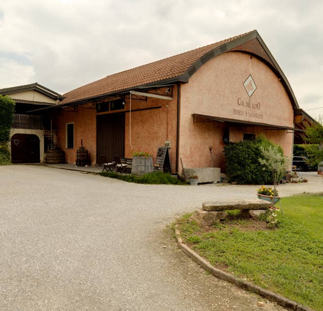 This round picture shows an Italian vineyard. You are standing on a gravelled car park in front of the production building. On the right-hand side is a stone bench and a few planted flowers. Behind it you can see the large, red-coloured building. A large wooden roller shutter door and the round clad roof ridge are striking features. The lettering "Col del Lupo" can be seen in small letters on the front of the building. 