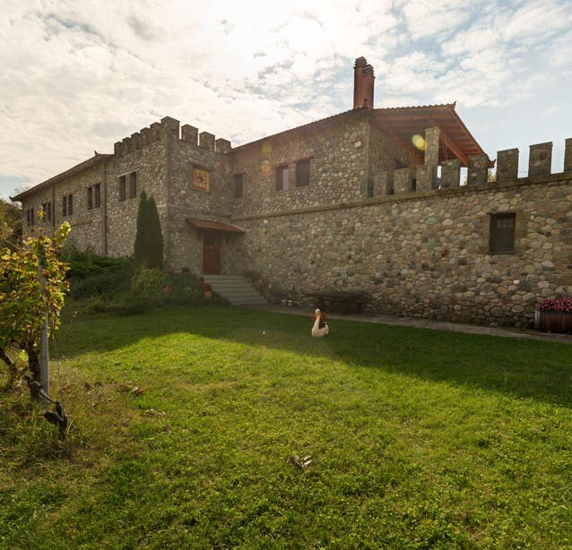 This round picture shows the Liakou winery in Greece in sunny weather. In the foreground, a short-cut lawn can be seen. A large stone building can be seen in the background. This building is made of rough, unworked natural stone. The battlements are striking, like those of a medieval castle.