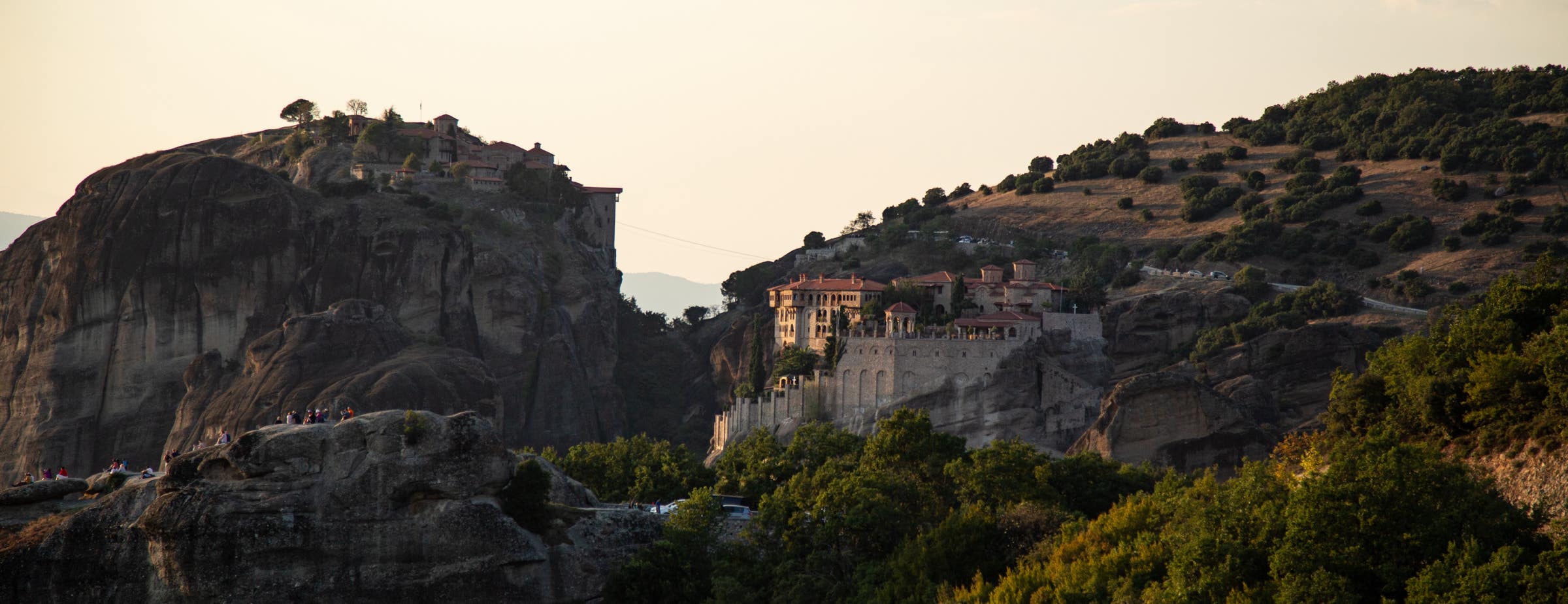 Slika prikazuje pogled na stijene i dva samostana Meteora. Lijevi samostan nalazi se na vrhu stijene, a desni samostan je izgrađen na stijenama na strani većeg brda.