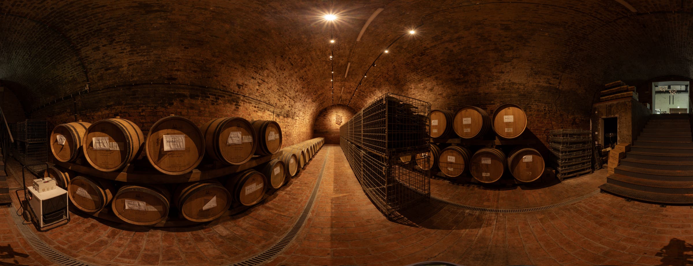 L'immagine mostra una vista a 360° di una cantina adibita allo stoccaggio delle botti lungo le pareti. Al centro della sala, lungo il corridoio, sono collocate cassette metalliche per la conservazione delle bottiglie di vino in vetro.